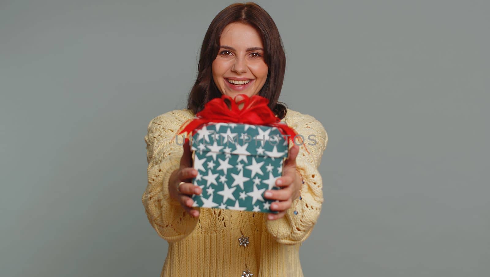 Positive smiling pretty woman presenting birthday gift box stretches out hands, offer wrapped present career bonus, celebrating party. Young adult girl isolated alone on gray studio background