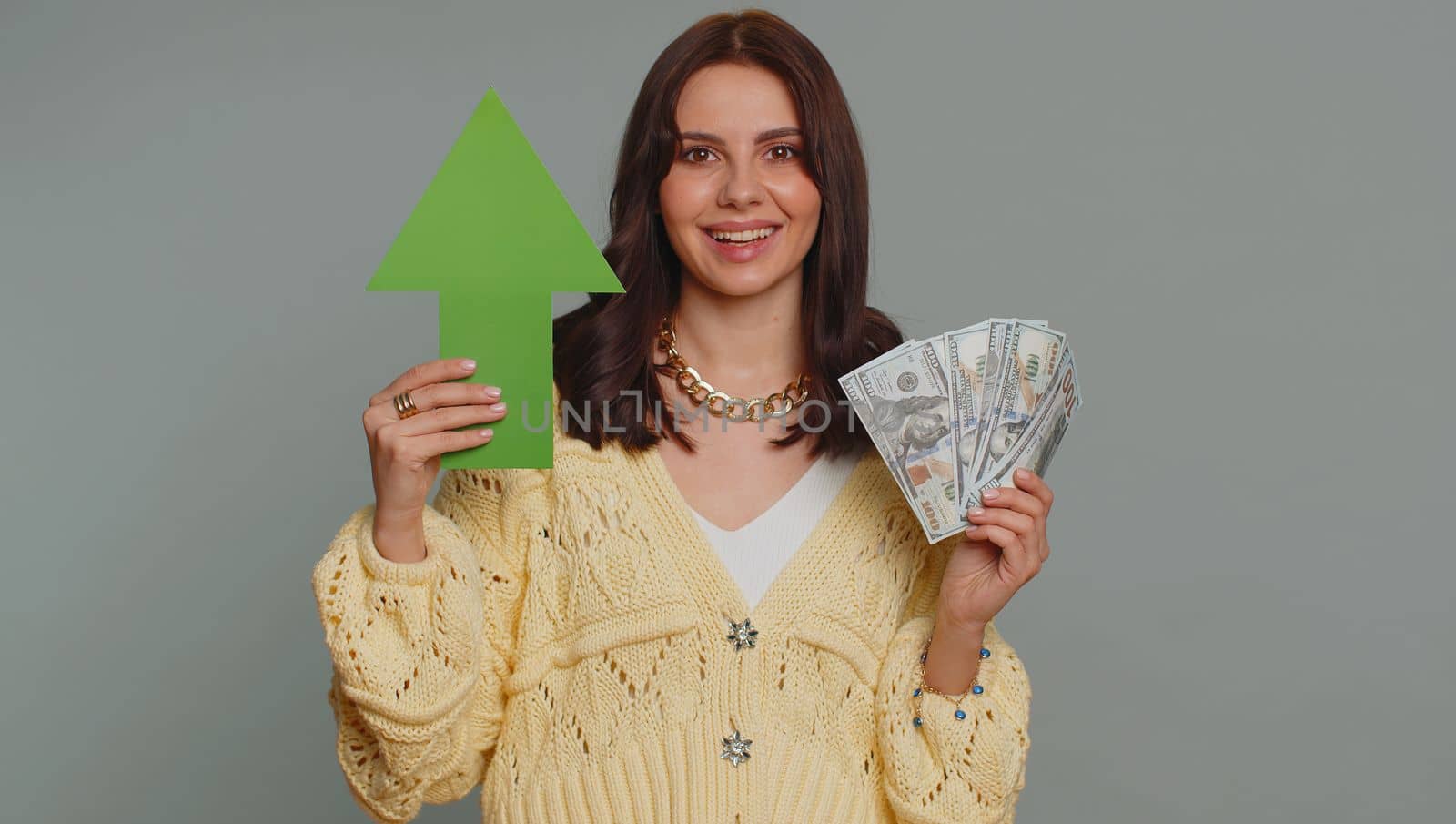 Happy pretty woman winner holding arrow sign pointing up, career growth and money dollar exchange increase. Young adult girl showing up indicating successful development progress on gray background