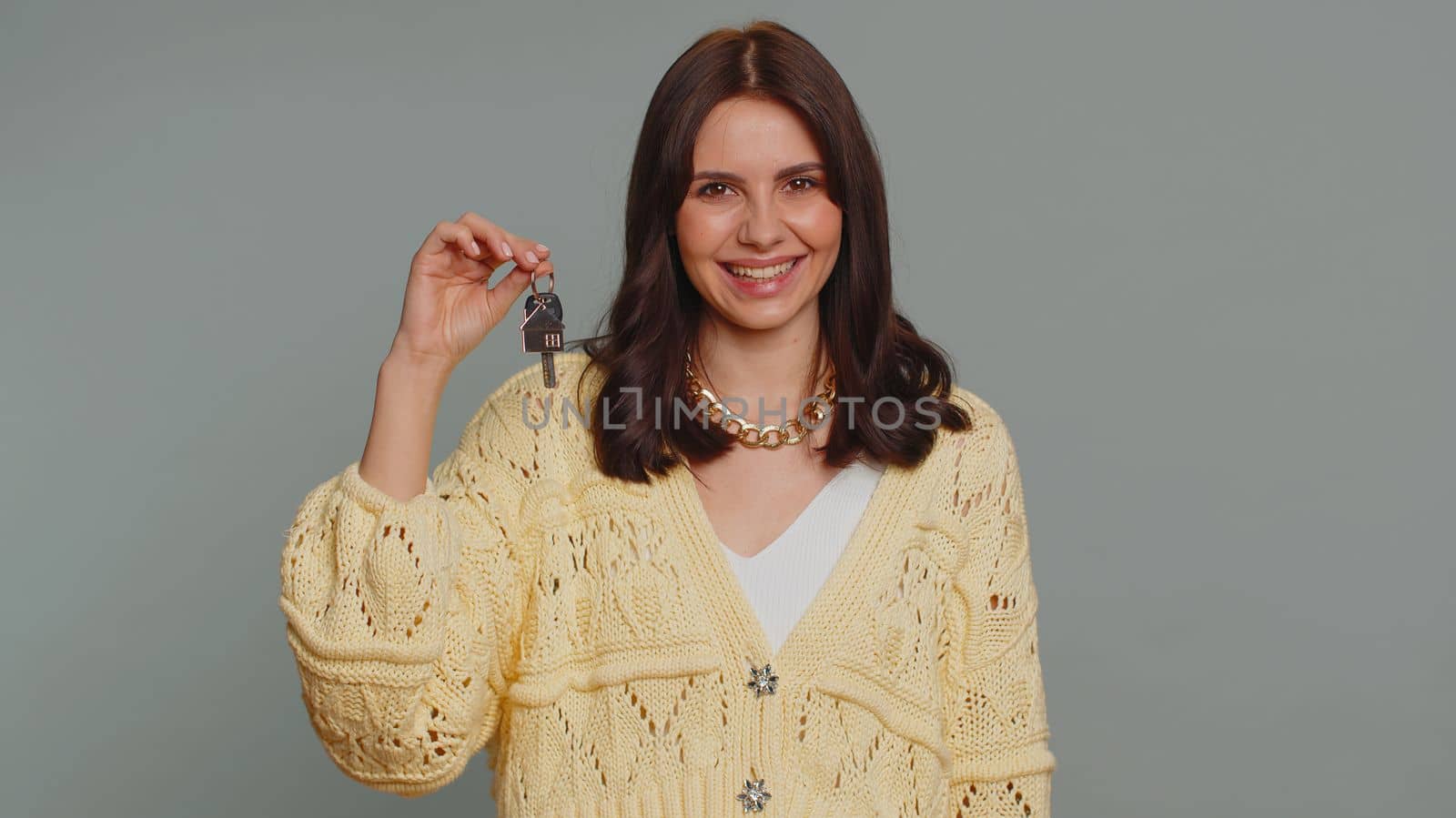 Pretty young woman in cardigan real estate agent lifting hand showing the keys of new home house apartment, buying, renting property, mortgage loan. Millennial girl isolated on gray studio background