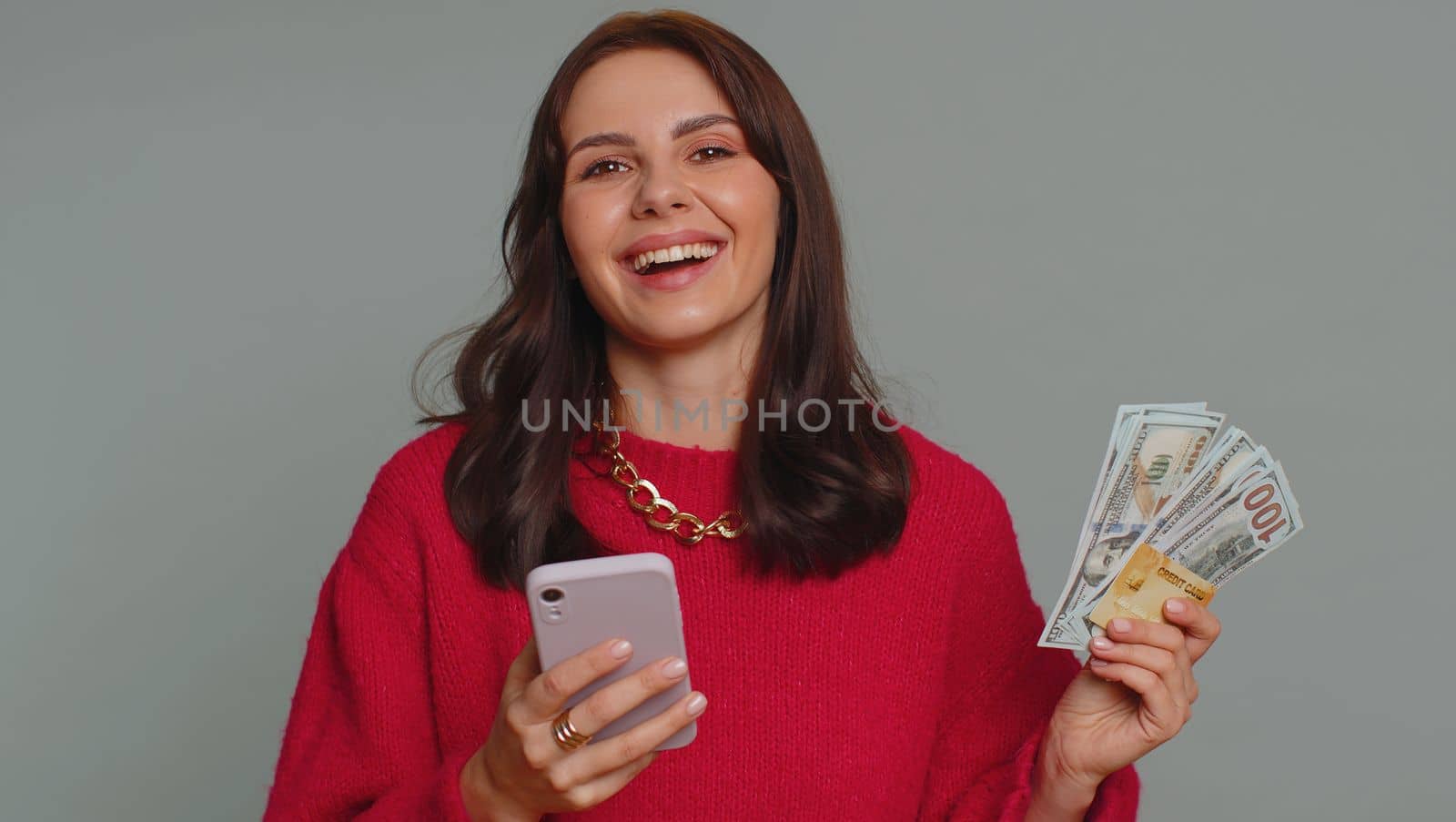 Sincere one woman customer using credit bank card and smartphone while transferring money, purchases online shopping, payment. Finance and internet. Young girl isolated alone on gray background