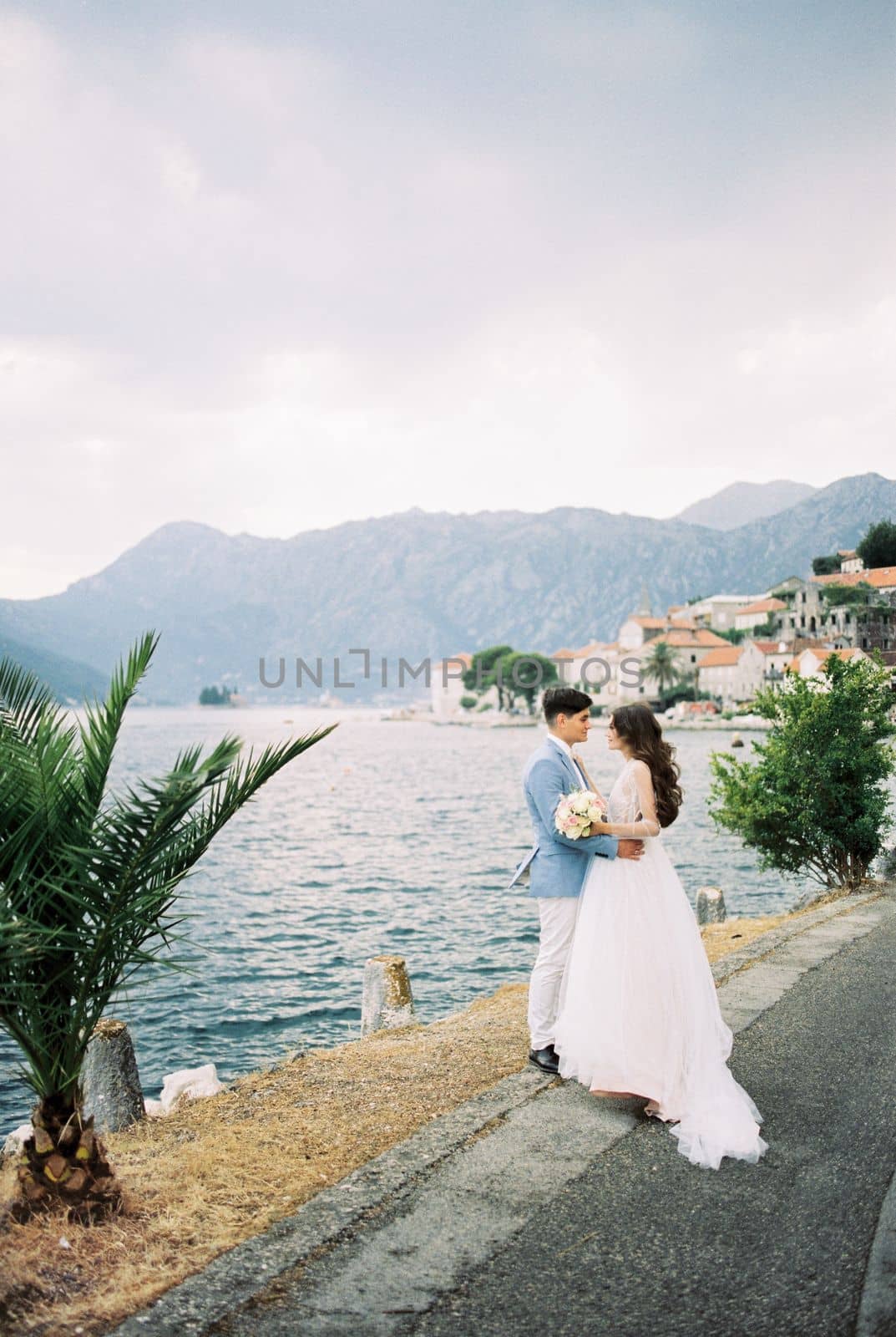 Bride and groom stand hugging on the embankment of Perast against the backdrop of the mountains. Montenegro. High quality photo