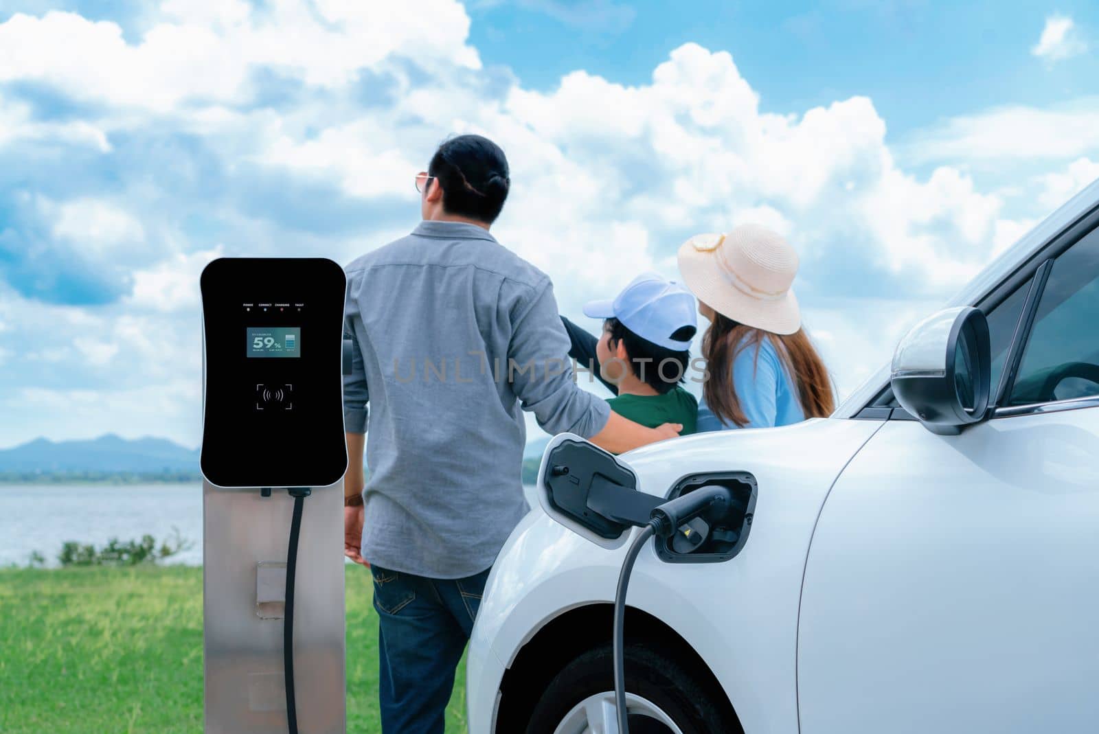 Concept of progressive happy family with electric vehicle enjoy their time at natural outdoor scenic with lake, greenfield and cloudscape background. Electric vehicle driven by green renewable energy.