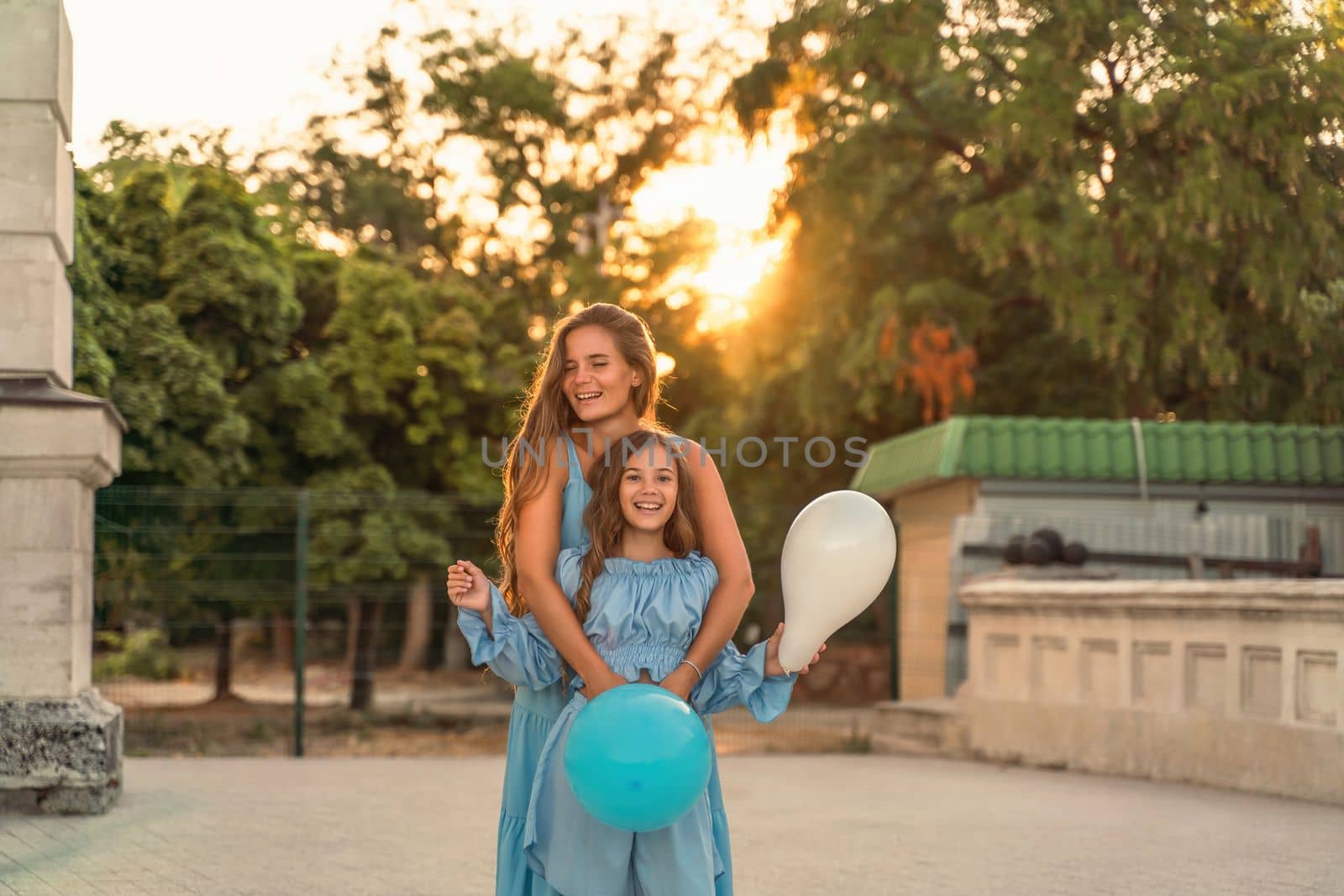 Mother daughter family sunset. Portrait of mother and daughter in blue dresses with flowing long hair against the backdrop of sunset. A woman hugs and presses the girl to her, holding balloons in her hands by Matiunina