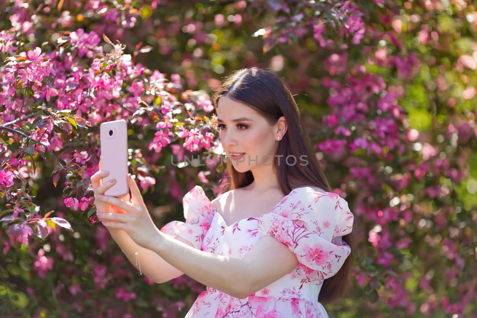 A beautiful brunette girl with long hair is standing in a pink blooming apple garden, on a sunny day, holding a pink smartphone. Close up