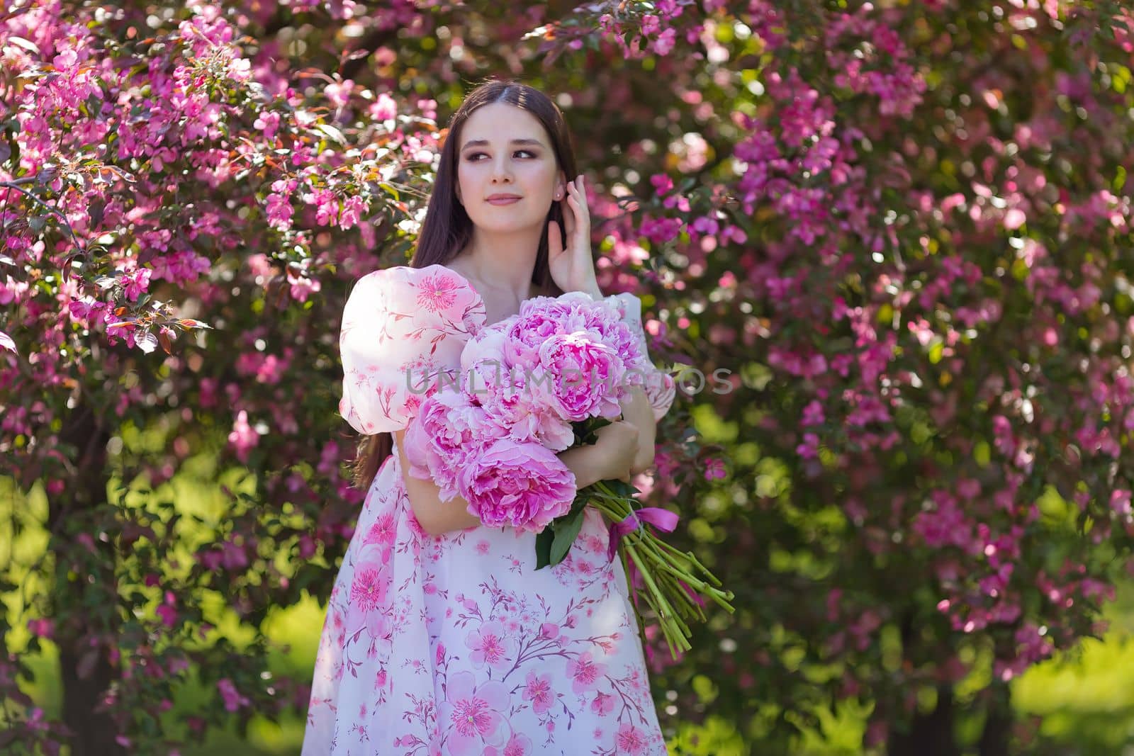 A portrait of pretty brunette in a light pink dress, with a large bouquet of pink peonies, stands near pink blooming apple trees, in summer in the garden on a sunny day. Copy space.