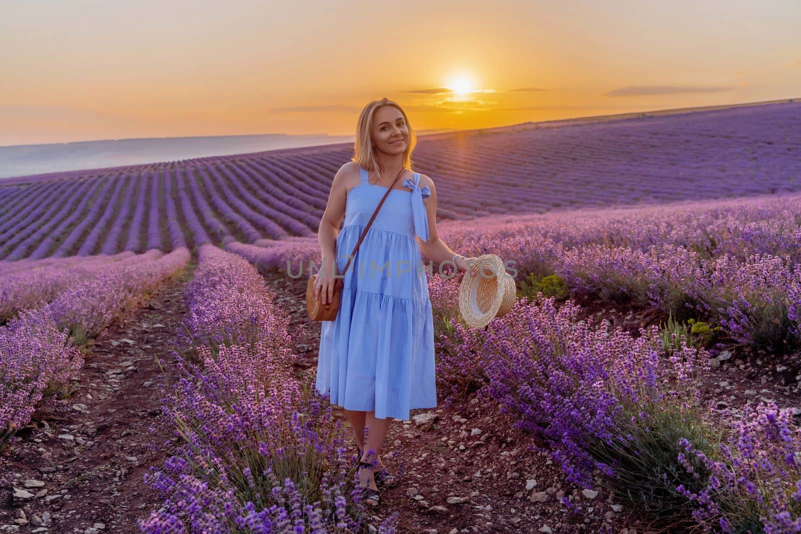 Woman lavender field sunset. Romantic woman walks through the lavender fields. illuminated by sunset sunlight. She is wearing a blue dress with a hat