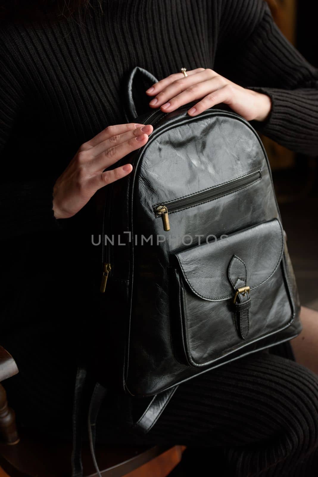 a brunette girl in a knitted black dress poses while sitting with a shiny black leather backpack in her hands