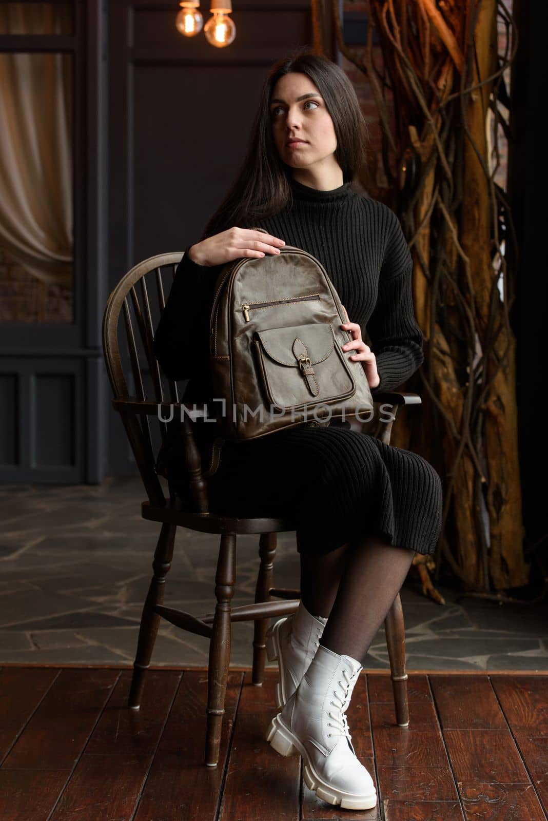 a brunette girl in a knitted black dress poses while sitting with a shiny brown leather backpack in her hands