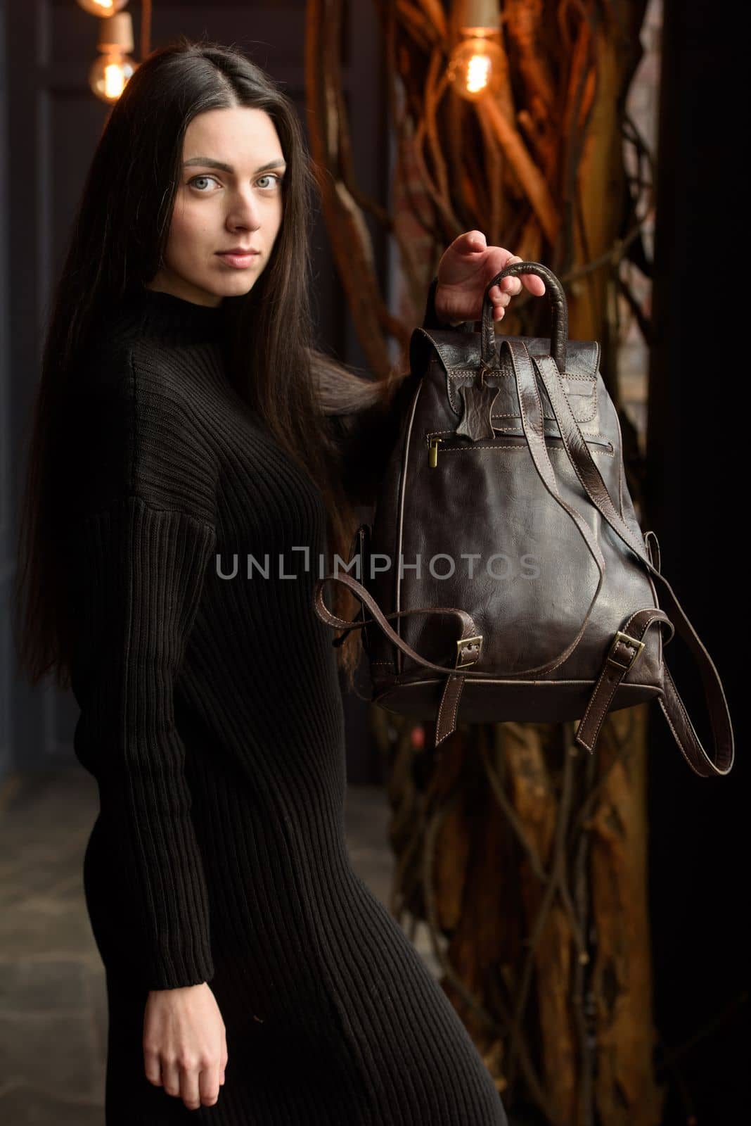 a brunette girl in a knitted black dress poses while standing with a shiny brown leather backpack in her hands