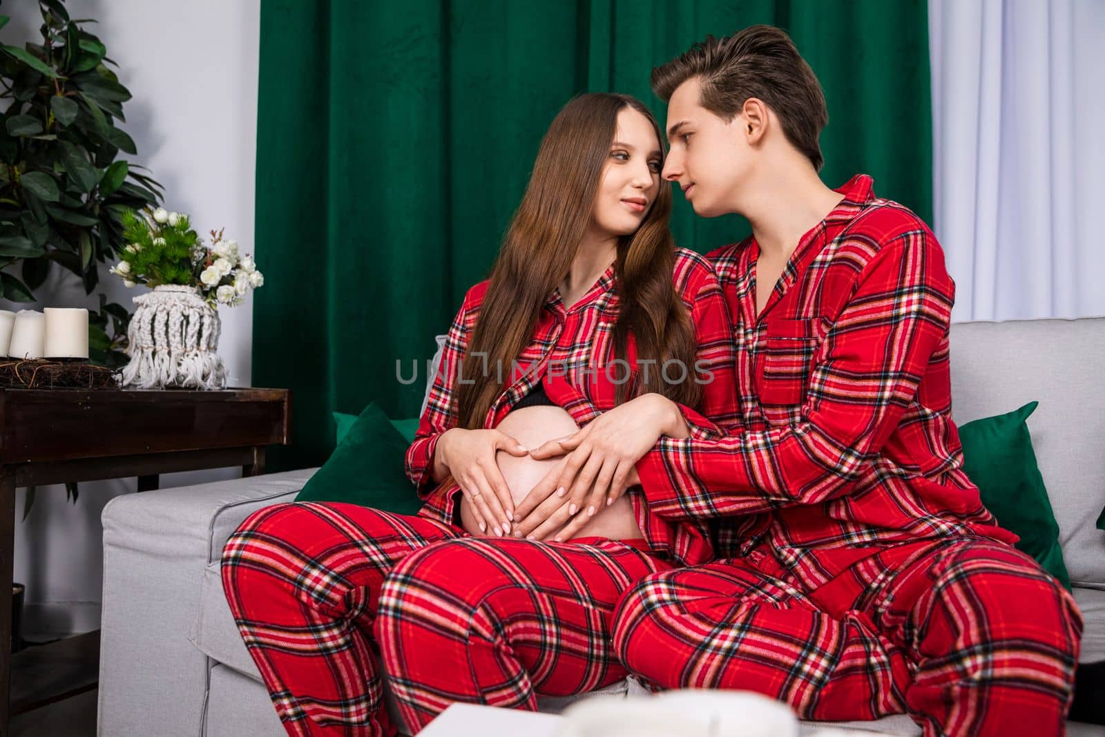 A boy and a girl sit on a light gray sofa and look deeply into each other's eyes. The setting is romantic and there are a lot of accessories in dark green, such as, pillows.