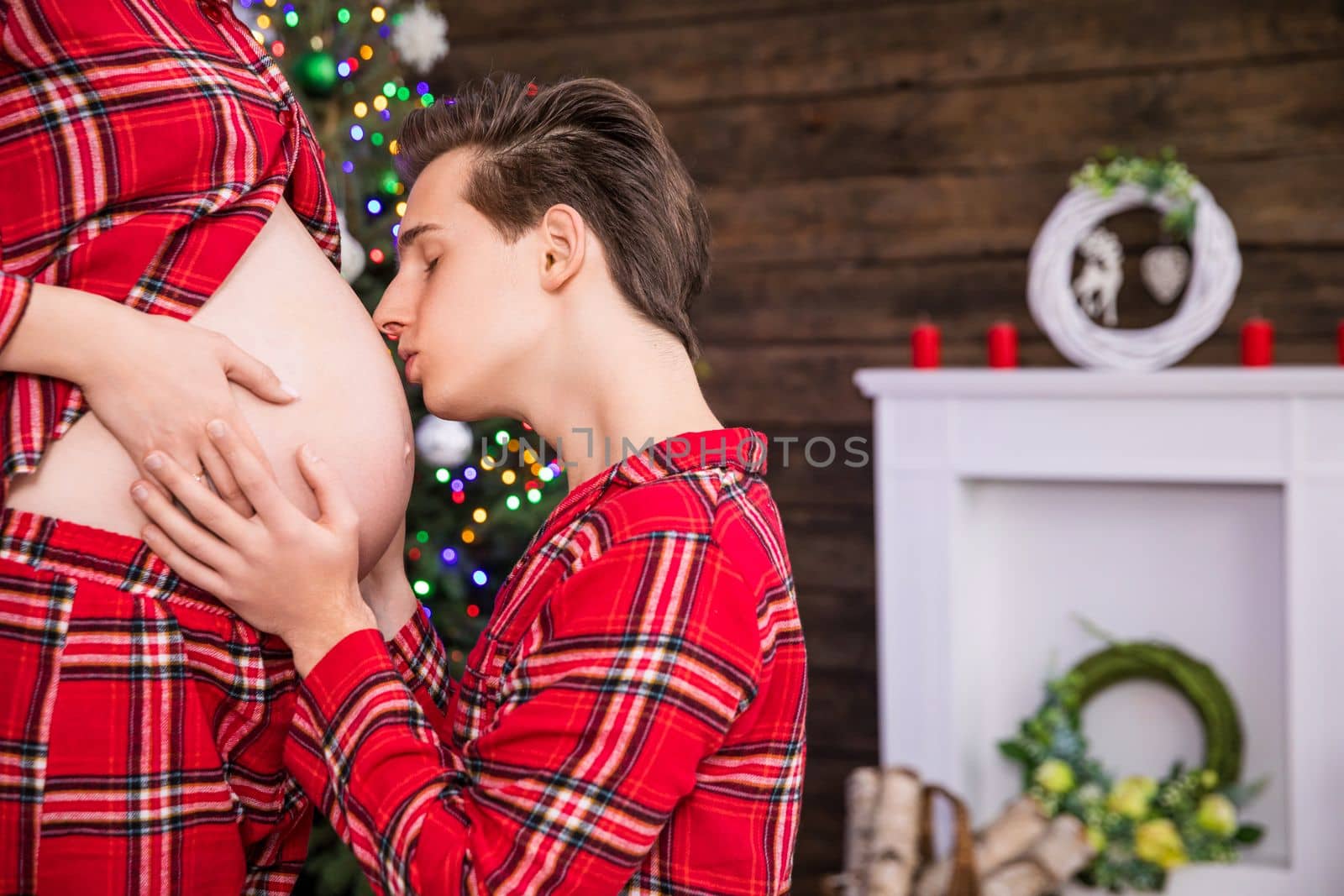 Against a background of Christmas decorations and a Christmas tree, a husband kneels in front of his wife. Close-up on the profile of the pregnant belly that the kneeling man is kissing. The partners hold hands on the pregnant belly.