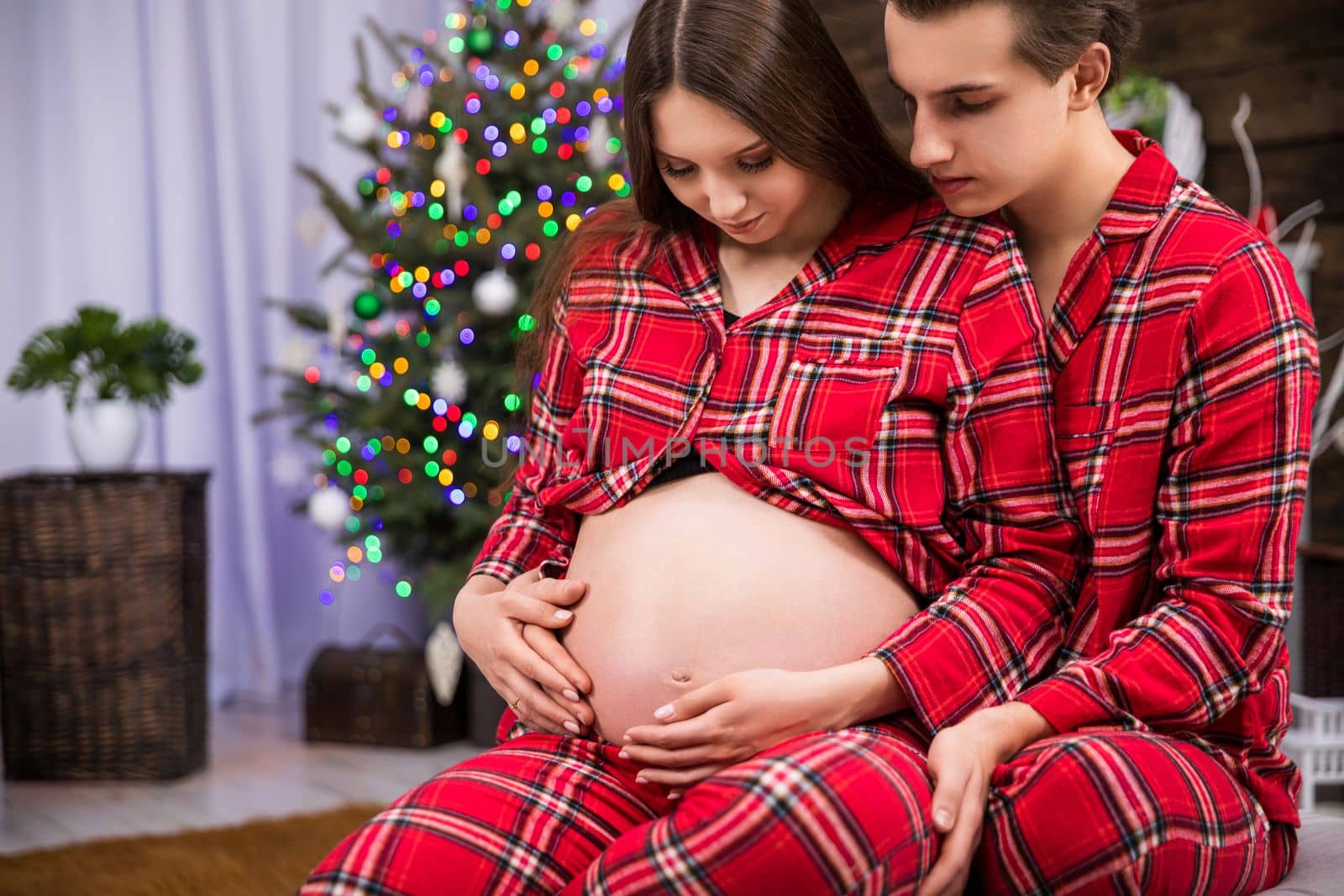 A couple expecting a baby sits hugging. The woman leans against the man with her back and exposes her pregnant belly. The man hugs the woman and strokes her pregnant belly.