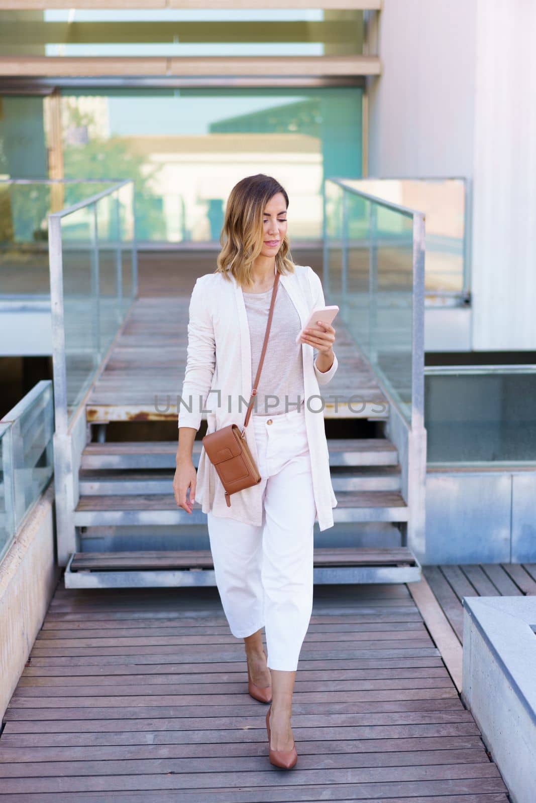 Full body woman in stylish clothes strolling on boardwalk and using mobile phone outside contemporary building