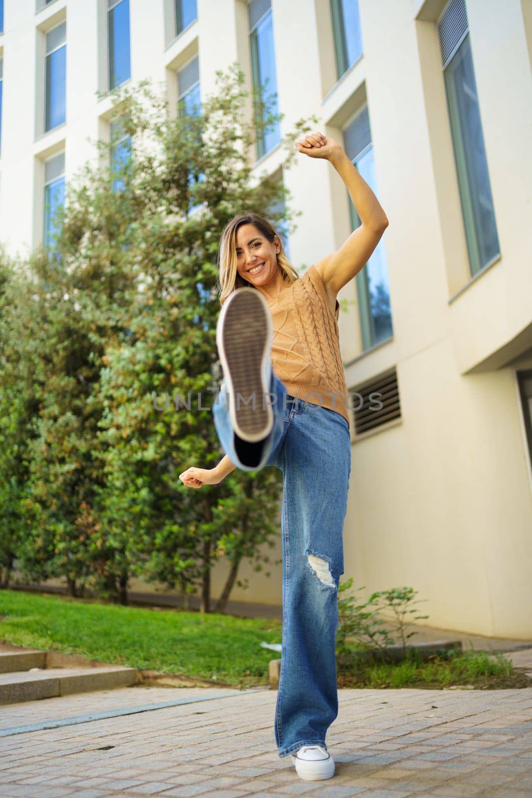 Full body cheerful adult woman in stylish outfit kicking air and looking at camera with smile while dancing on city street