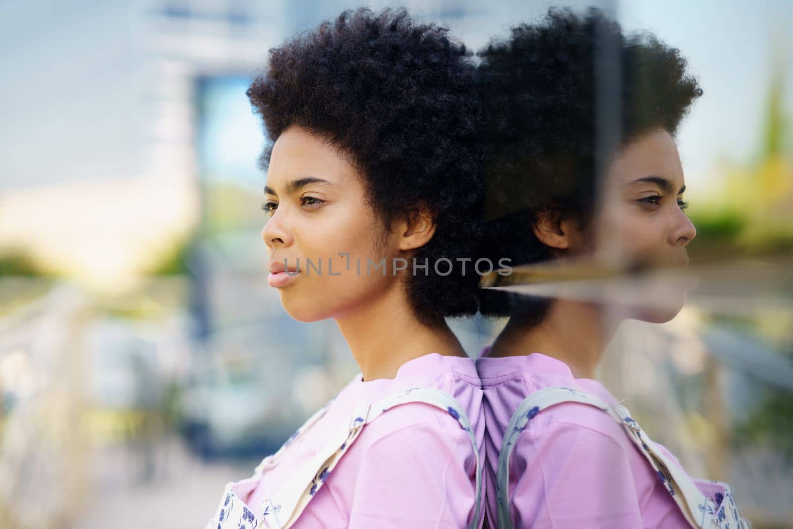 Side view of serious African American female with Afro hairstyle standing near glass house on street against blurred background in city