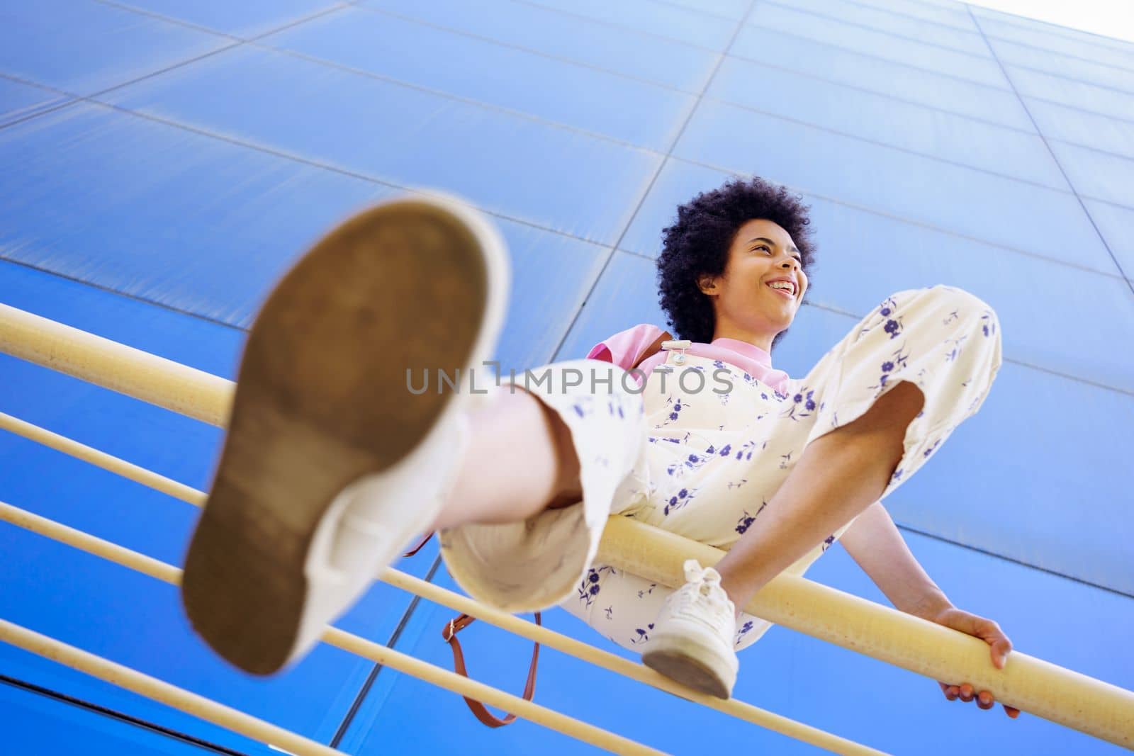 Low angle full body of African American female in overall looking away while sitting on railing near modern building in city