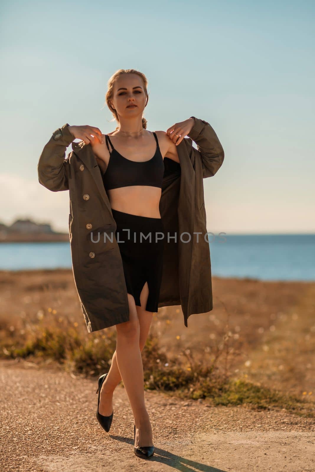 Portrait blonde sea cape. A calm young blonde in an unbuttoned khaki raincoat walks along the seashore, under a raincoat a black skirt and top.