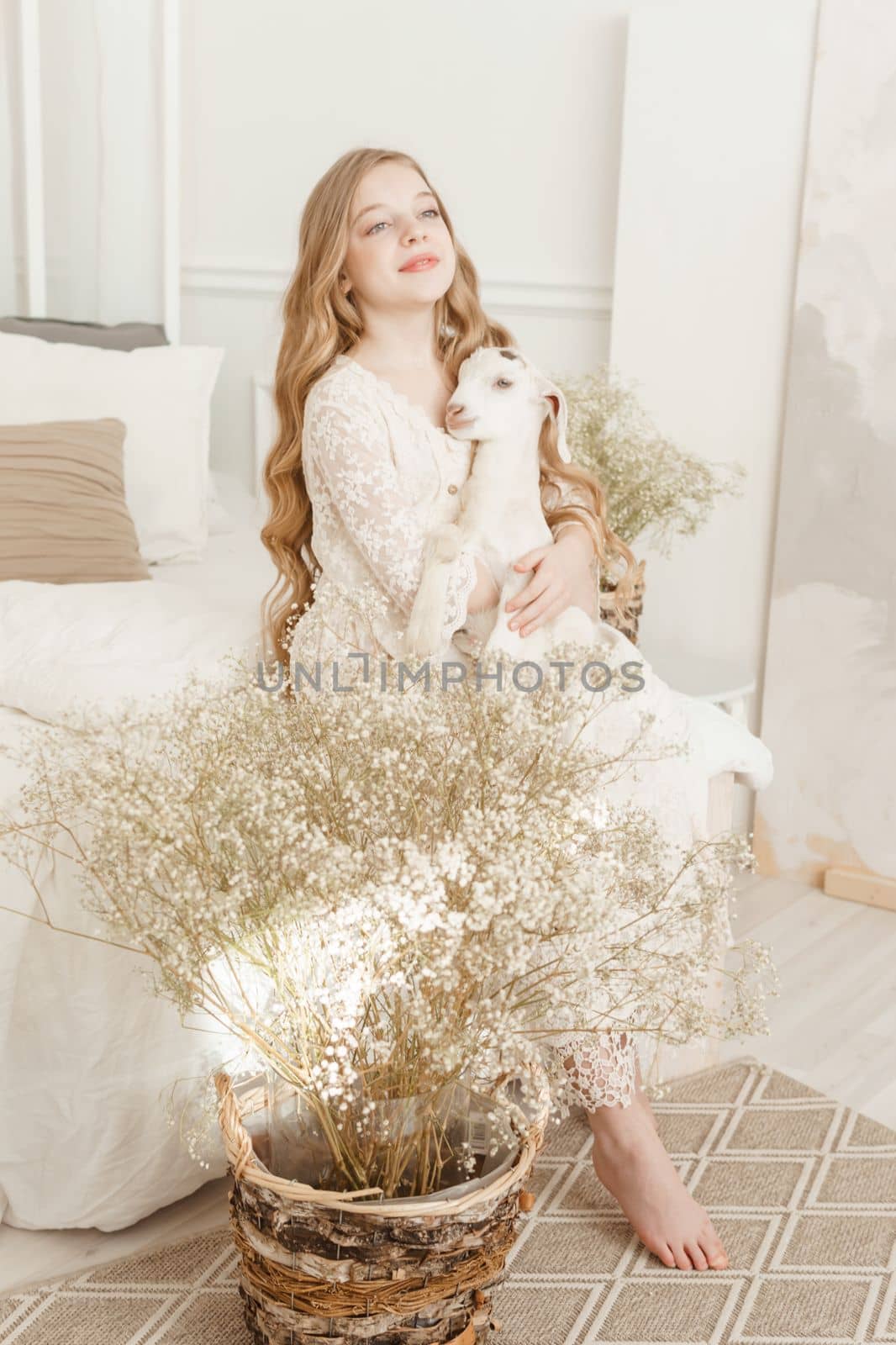 A blonde girl with long hair is sitting on the couch with a goat at home. The concept of animal care