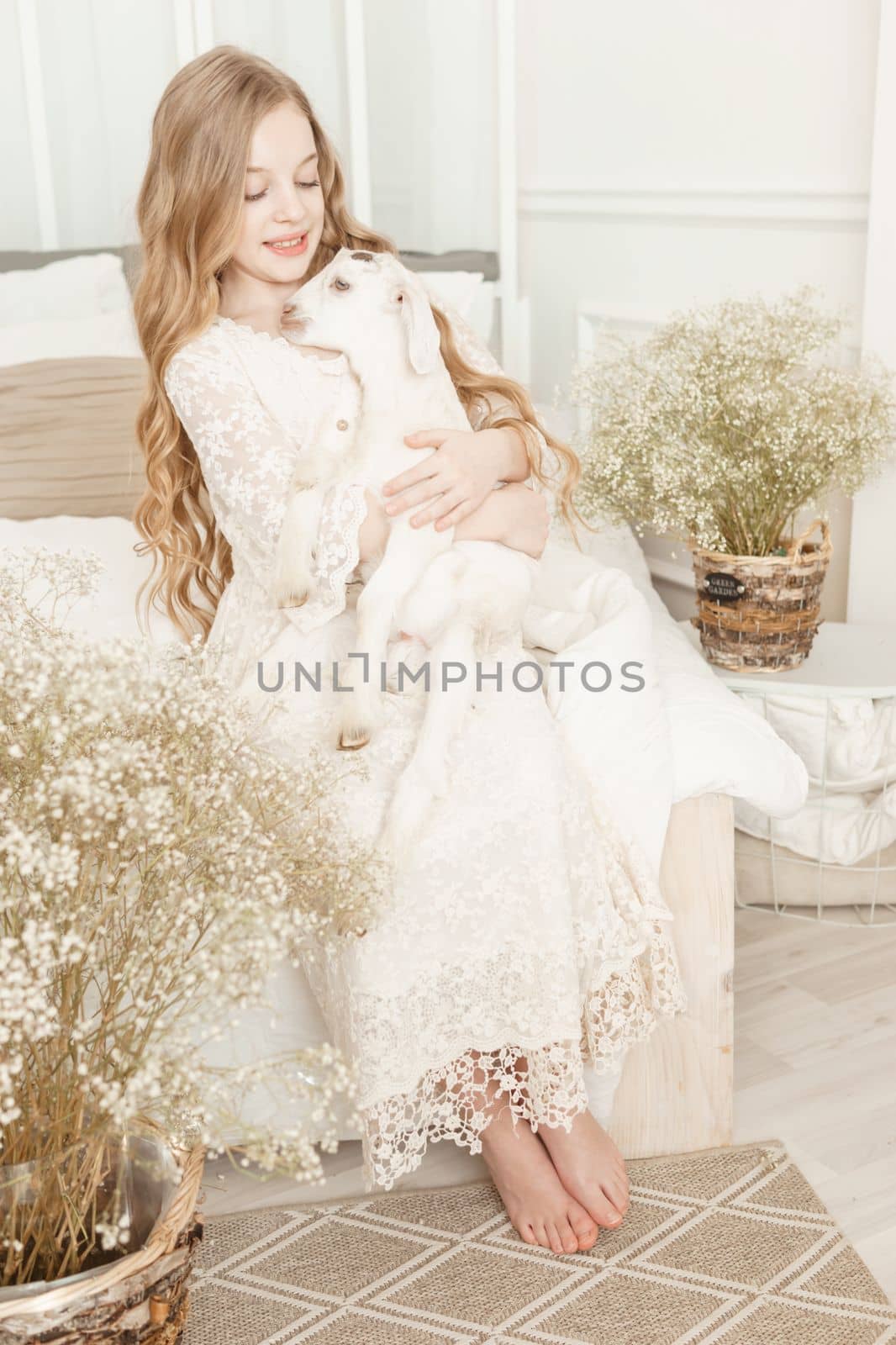 A blonde girl with long hair is sitting on the couch with a goat at home. The concept of animal care