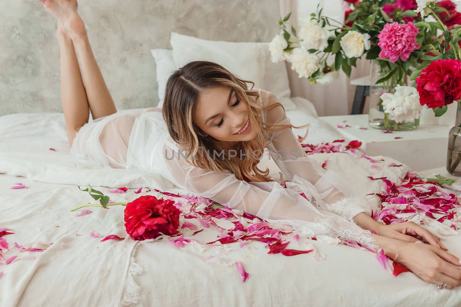 A beautiful girl in a white nightgown at home, surrounded by spring flowers on a large bed. A room decorated with bouquets of peonies.