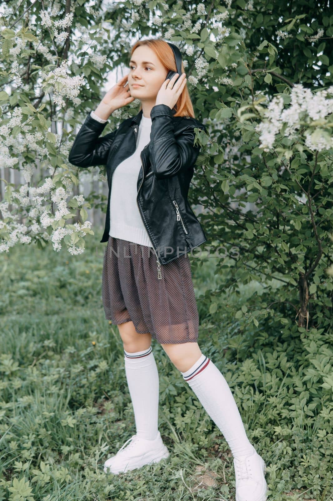 A red-haired teenage girl listens to music in headphones, configured via the phone. The time of spring flowering of cherry orchards.