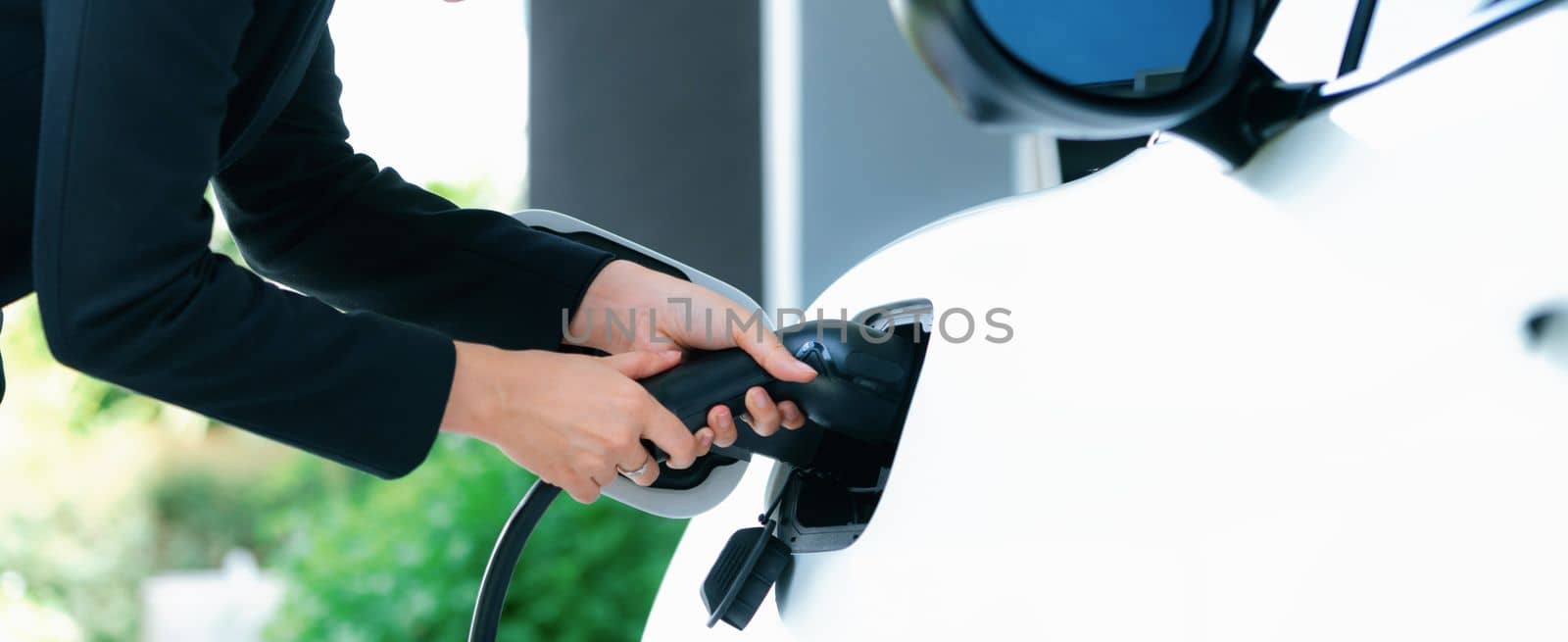 Closeup progressive woman install cable plug to her electric car with home charging station. Concept of the use of electric vehicles in a progressive lifestyle contributes to clean environment.