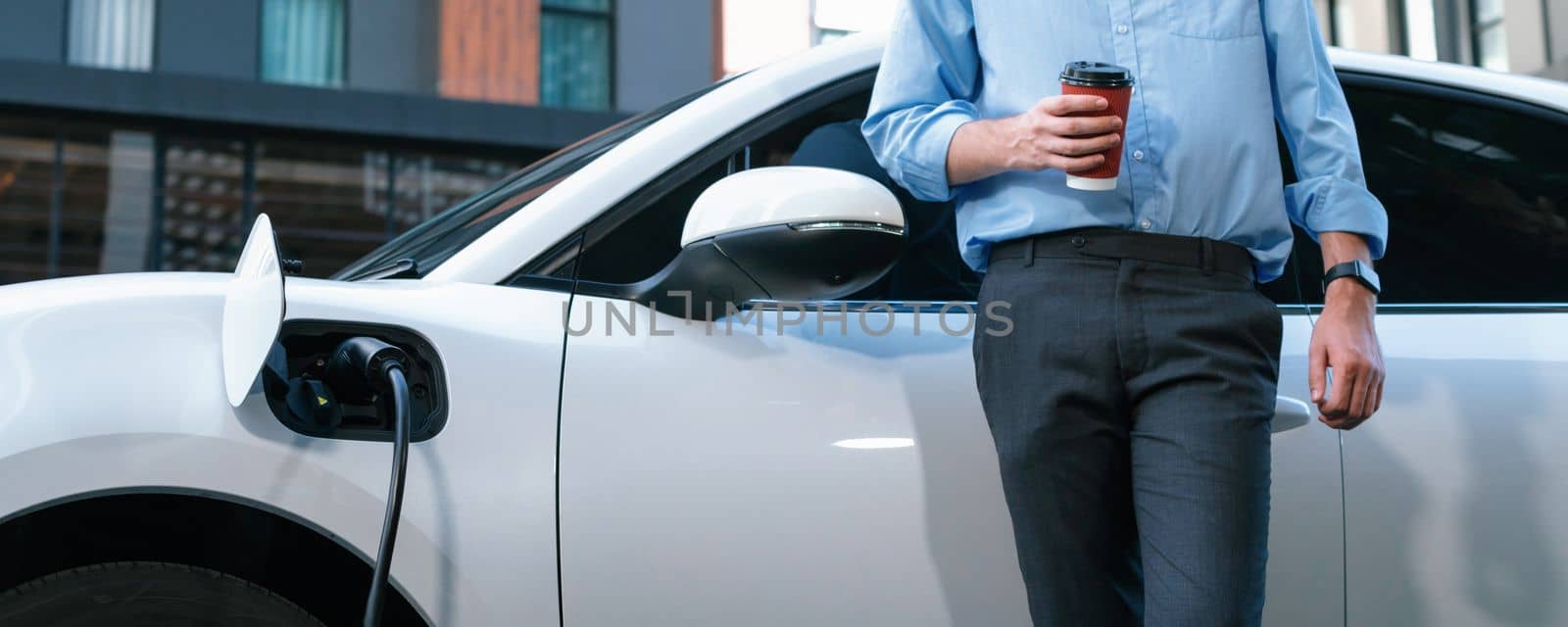 Progressive eco-friendly concept of parking EV car at public electric-powered charging station in city with blur background of businessman leaning on recharging-electric vehicle with coffee.