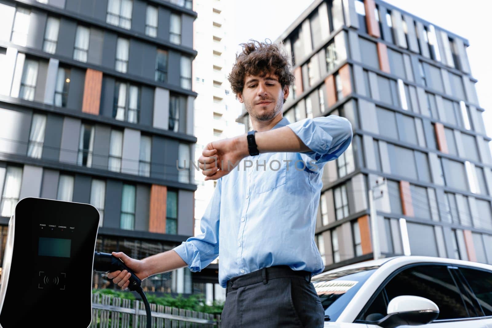 Businessman with smartwatch at modern charging station for electric vehicle with background of residential buildings as concept for progressive lifestyle of using eco-friendly as alternative energy.