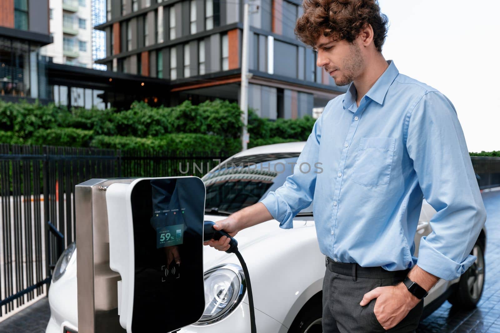 Progressive businessman insert charger plug from charging station to his electric vehicle with apartment condo building in background. Eco friendly rechargeable car powered by sustainable energy.