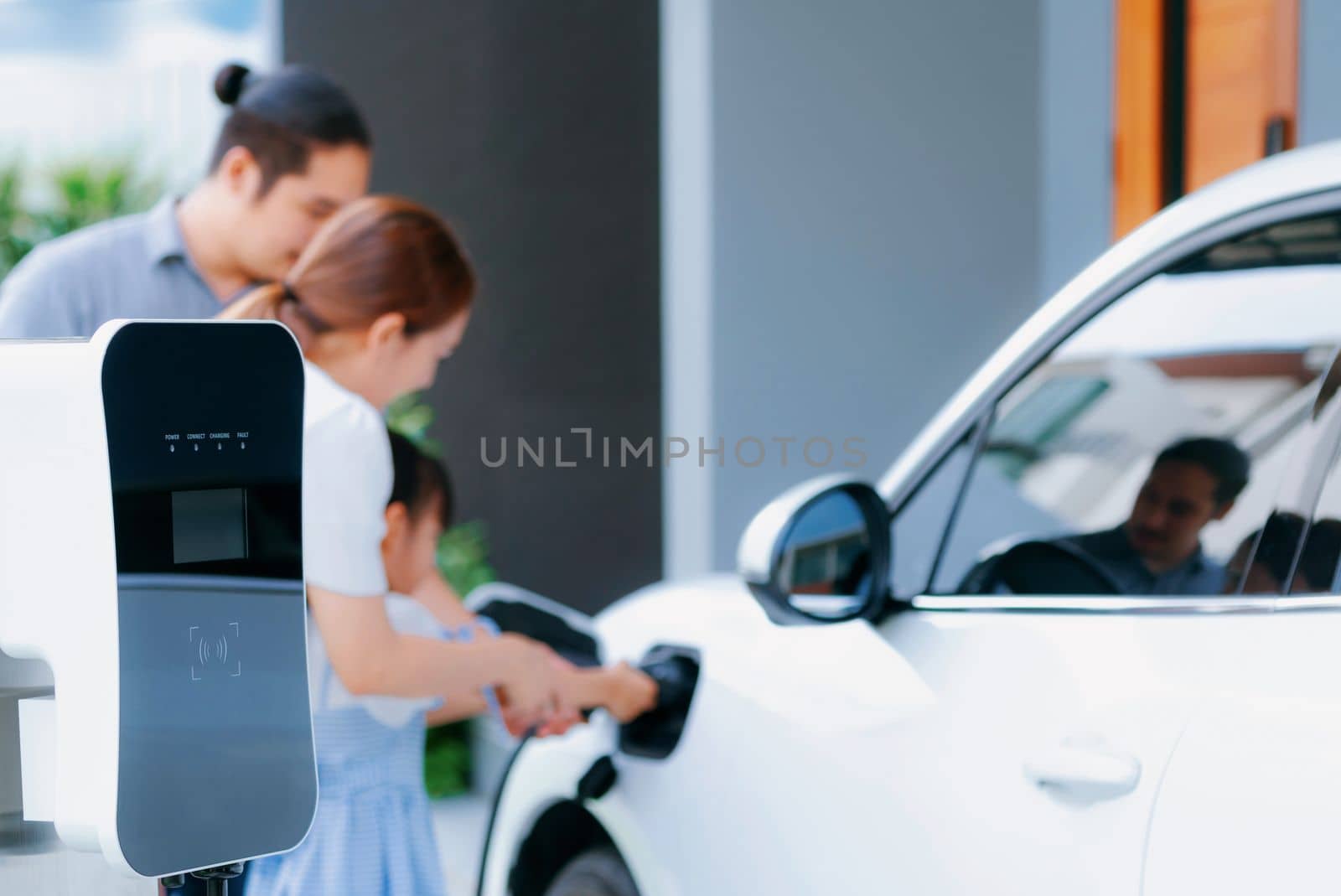 Closeup focus hand insert EV charger plug to electric vehicle at home charging station with blurred family in background. Progressive concept of green and clean energy to reduce CO2 emission by EV car