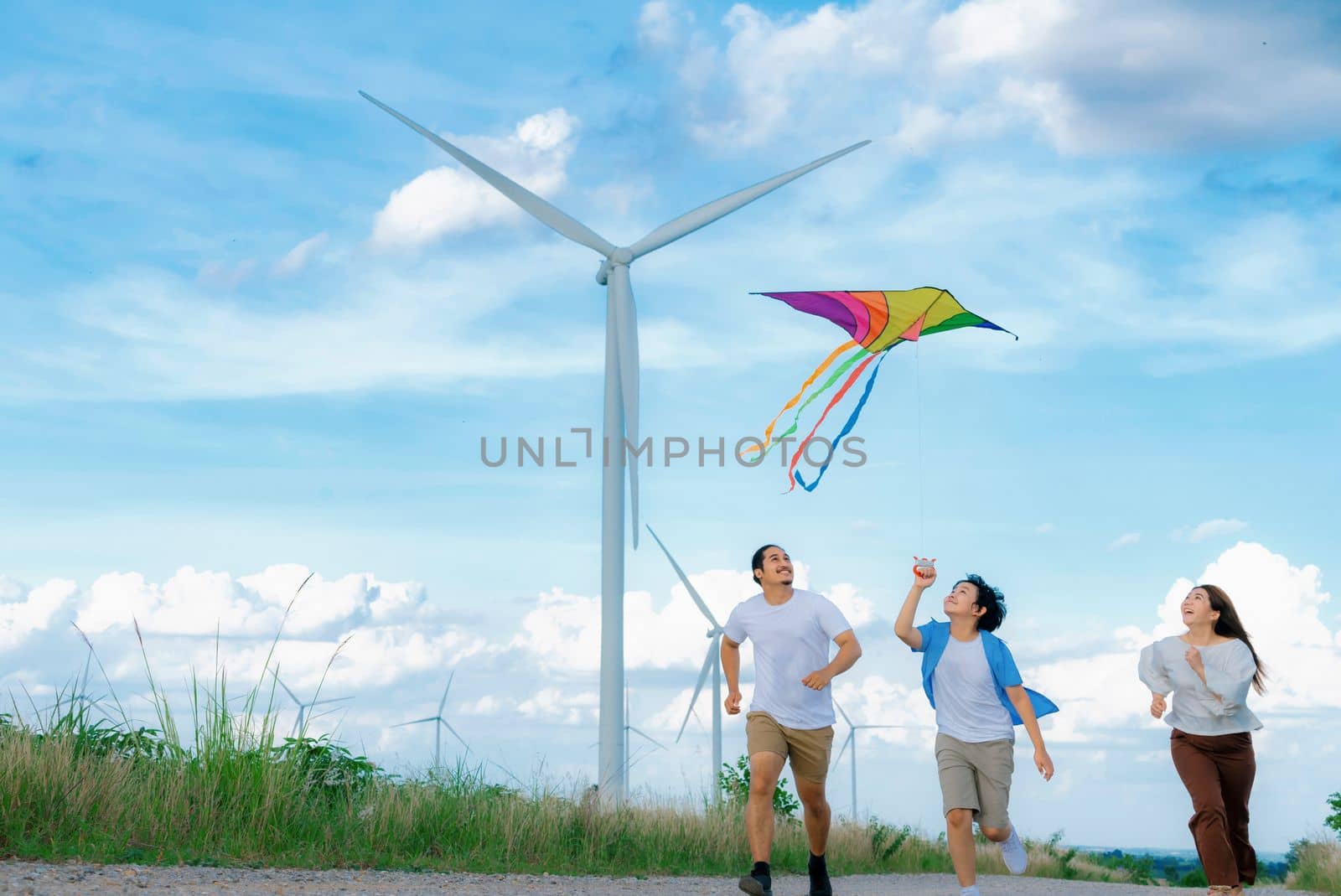 Progressive happy carefree family vacation concept. Young parents mother father and son run along and flying kite together on road with natural scenic on mountain and wind turbine background.