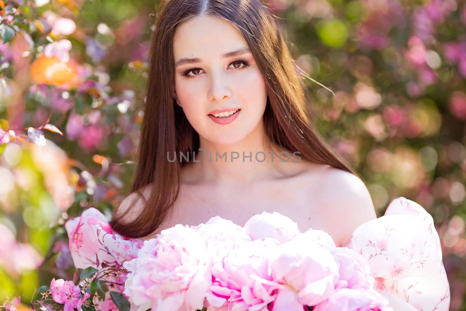 Close up of a smiling beautiful girl with a large bouquet of pink peonies , stands in a garden on a sunny day, look at camera. Copy space