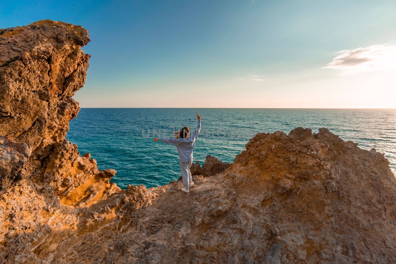 The tourist, standing with her hands raised, reaches the edge of the earth and sees a rock. The girl welcomes the sun. Design concept. A successful woman looks at the sunset with open arms. by Matiunina