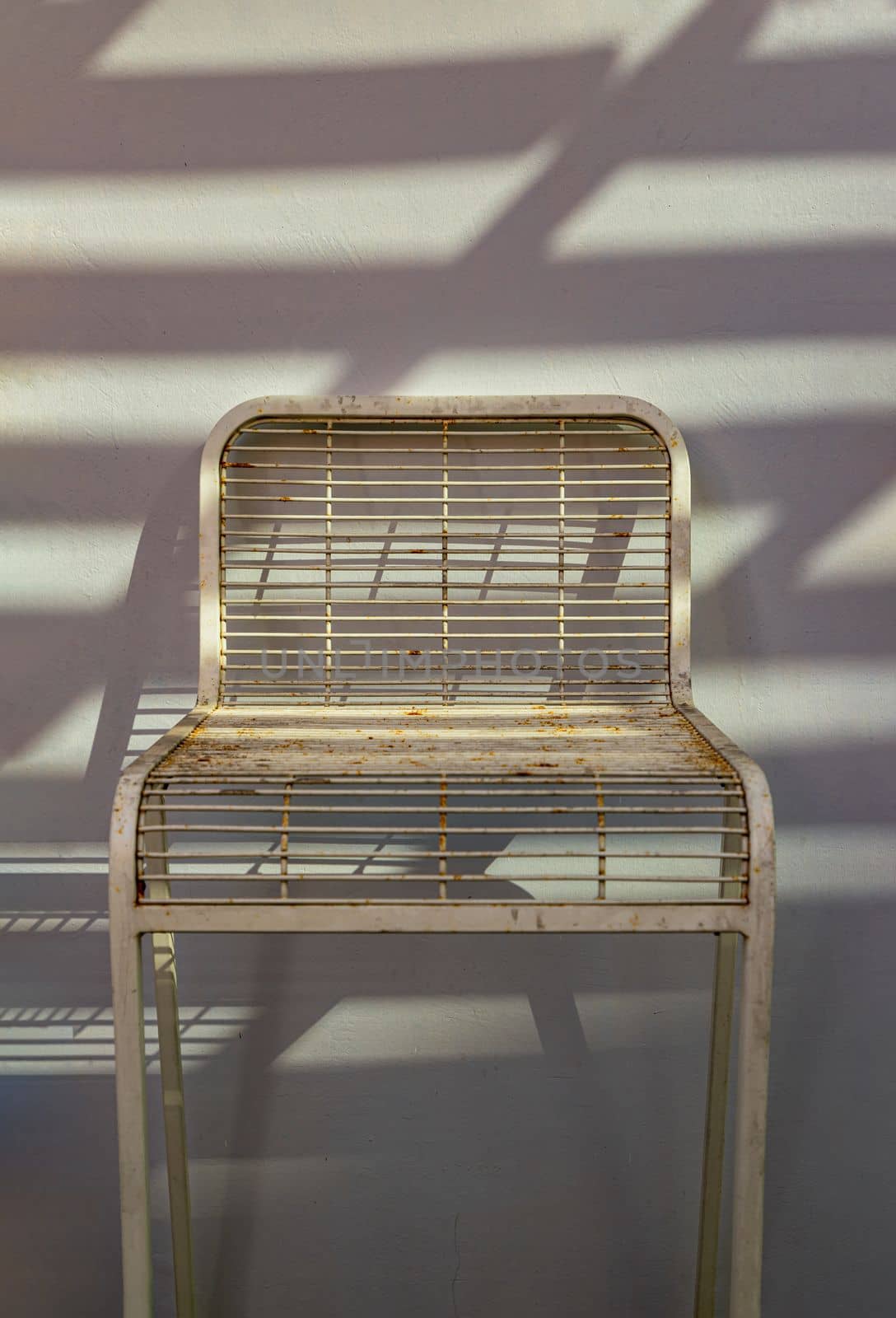 The white metal chair is stand alone in front of the white wall with sunlight shine through. Space for text, Selective focus.