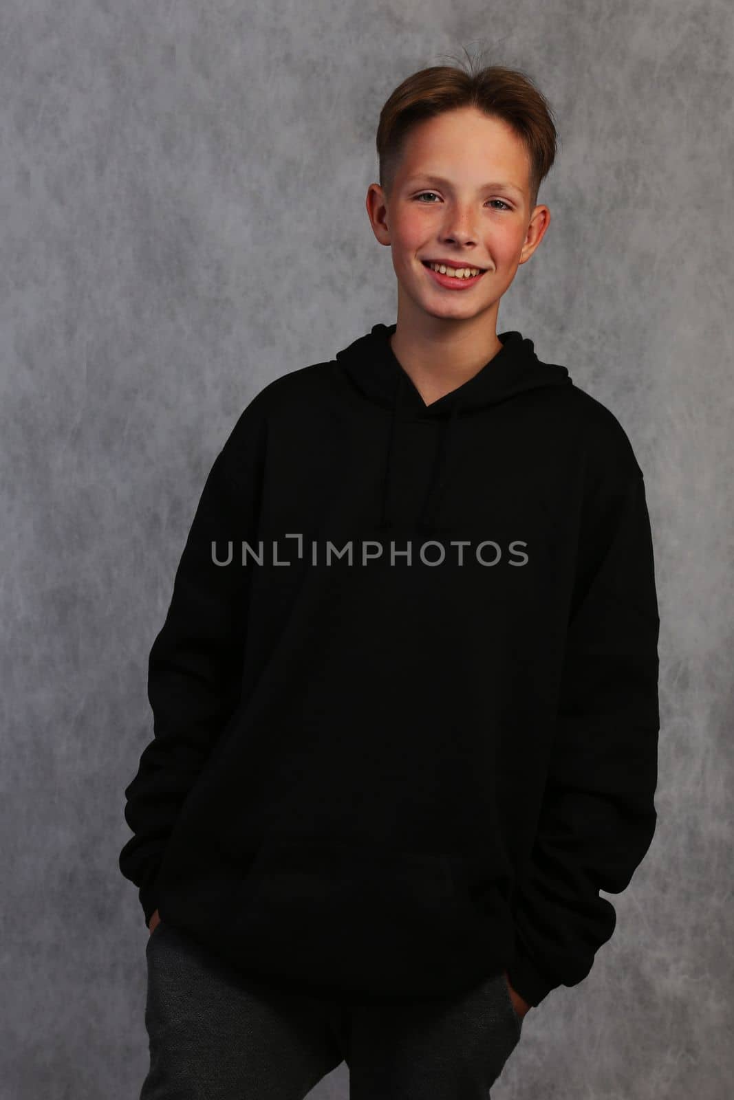 The boy poses holding his hands in his pockets. A boy in a sweatshirt is posing and smiling on a gray background.