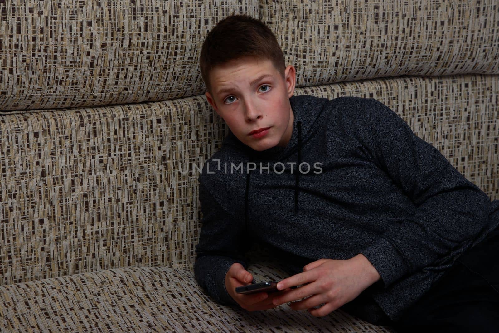 A boy in a sweatshirt lying on the sofa with a smartphone.