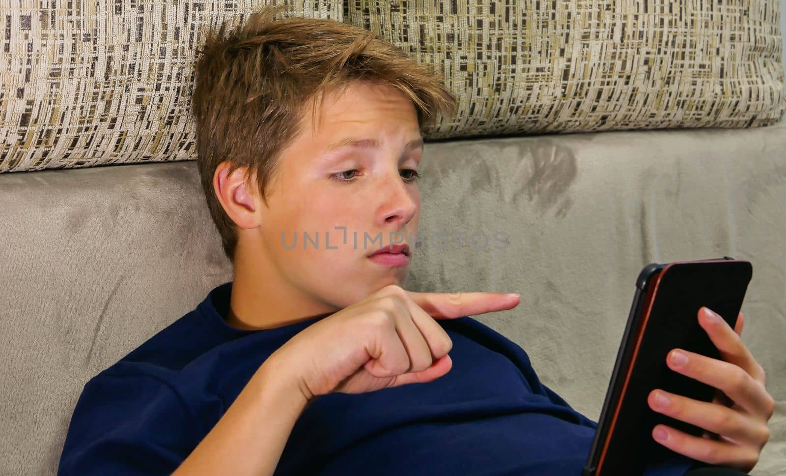 A handsome serious boy in a blue shirt sits on the sofa and reads an e-book. Teenager at home during self-isolation.