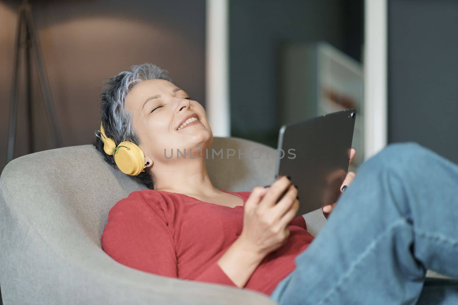Mature woman with grey hair is depicted listening to music while sitting on sofa in her home. She has her eyes closed and is holding a tablet in her hands. High quality photo