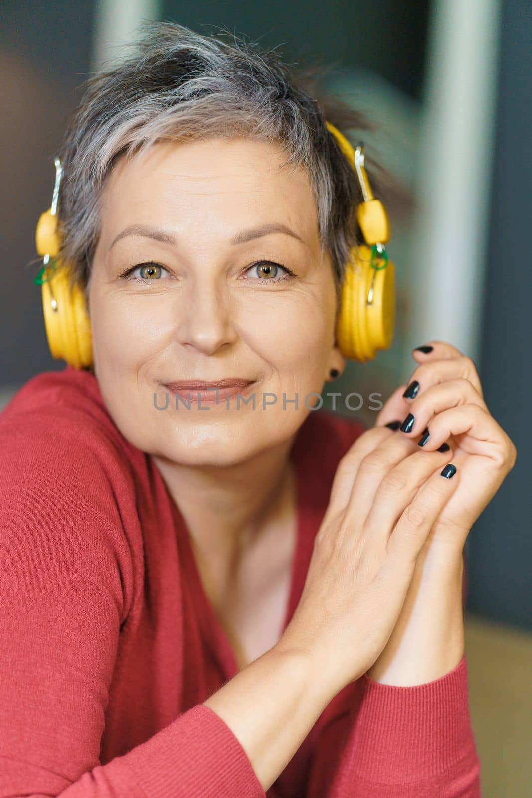 Close-up vertical portrait of a senior woman with grey hair and a pink blouse, smiling at the camera while wearing yellow headphones in her home. by LipikStockMedia