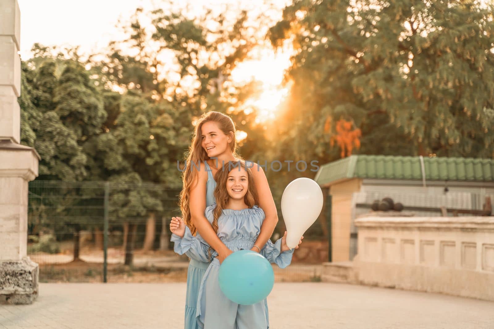 Mother daughter sunset. in blue dresses with flowing long hair against the backdrop of sunset. The woman hugs and presses the girl to her. They are looking at the camera