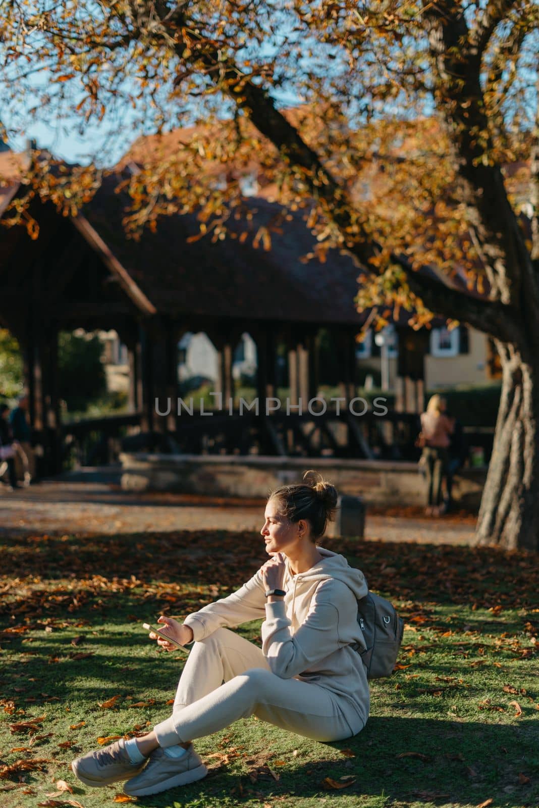 Young fashionable teenage girl with smartphone in park in autumn sitting at smiling. Trendy young woman in fall in park texting. Retouched, vibrant colors. Beautiful blonde teenage girl wearing casual modern autumn outfit sitting in park in autumn. Retouched, vibrant colors, brownish tones.