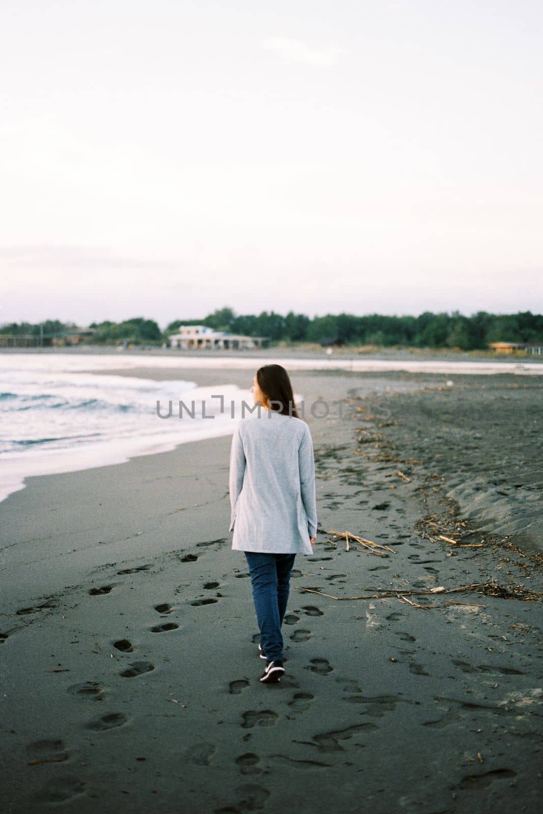 Young woman in a sweater walks along the beach and looks at the surf. High quality photo