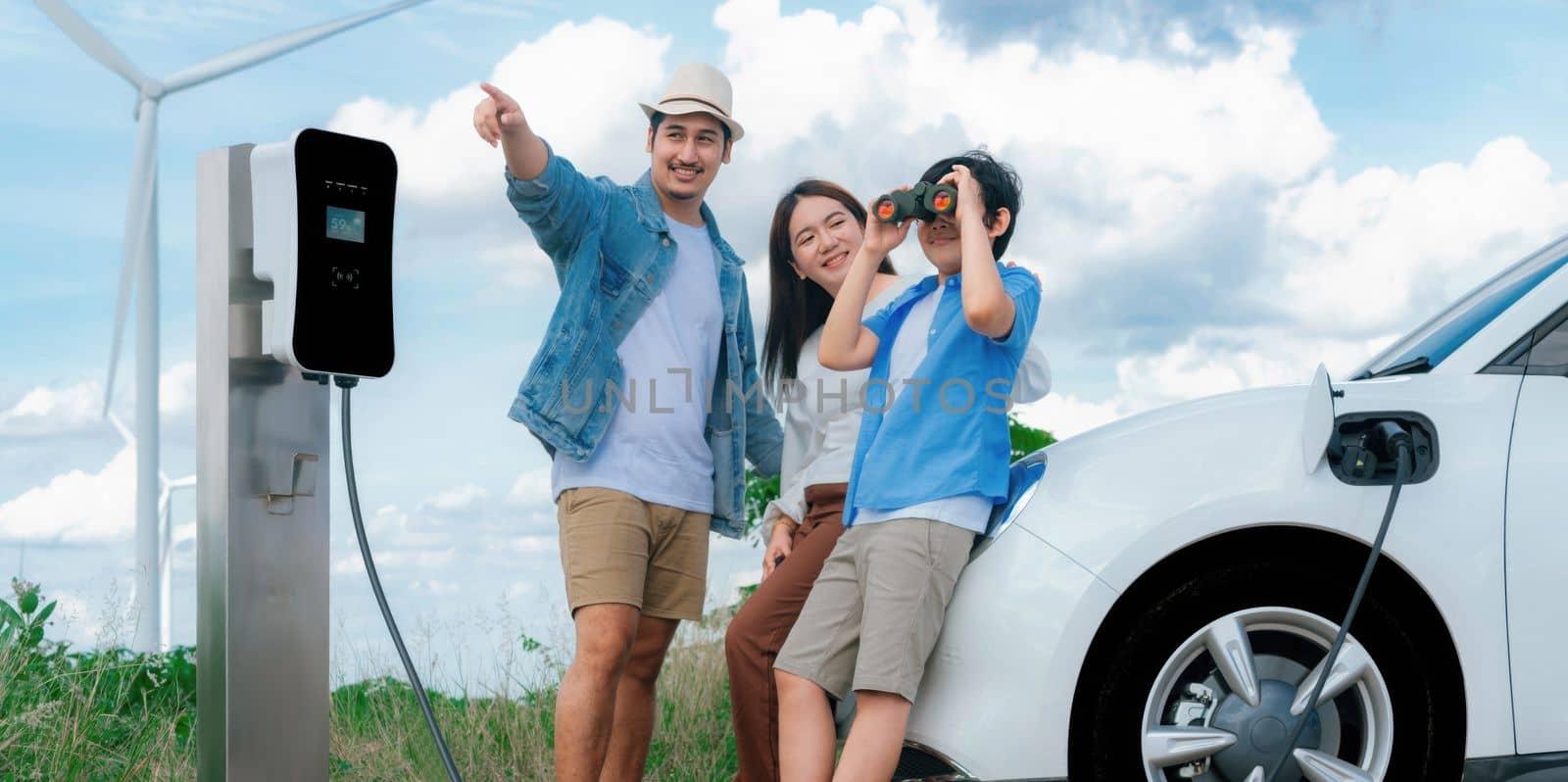 Concept of progressive happy family enjoying their time at wind farm with electric vehicle. Electric vehicle driven by clean renewable energy from wind turbine generator for charging station.