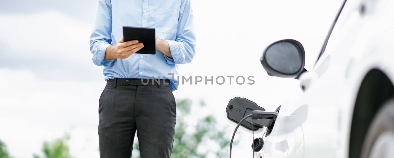 Closeup progressive suit-clad businessman with his electric vehicle recharge his car on public charging station in modern city with power cable plug and renewable energy-powered electric vehicle.