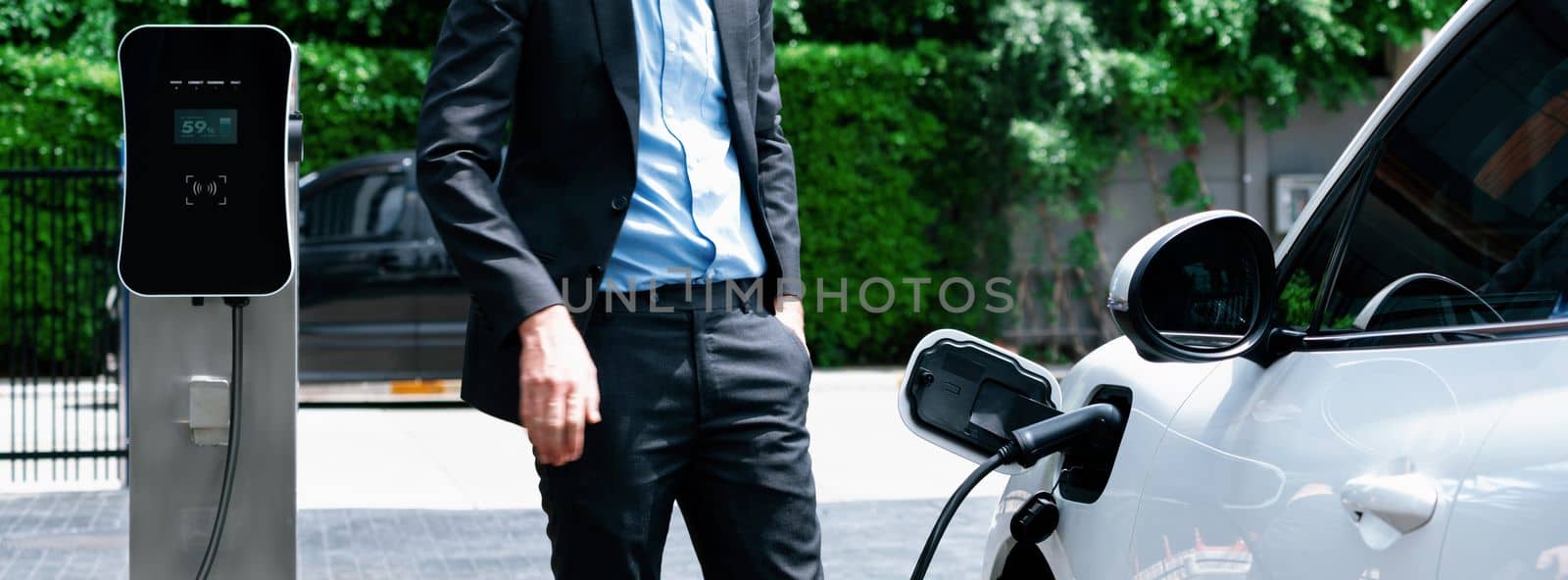 Closeup progressive suit-clad businessman with his electric vehicle recharge his car on public charging station in modern city with power cable plug and renewable energy-powered electric vehicle.