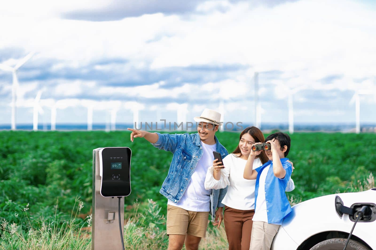 Concept of progressive happy family enjoying their time at wind farm with electric vehicle. Electric vehicle driven by clean renewable energy from wind turbine generator for charging station.