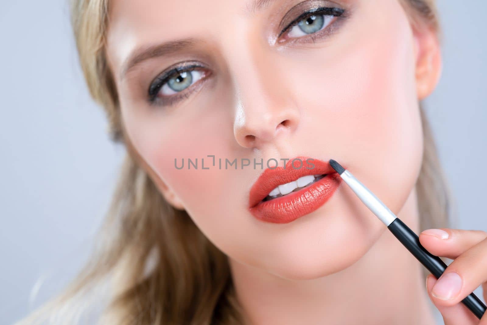 Closeup beautiful young woman with flawless healthy skin and natural makeup putting alluring fashion glossy red lipstick on her lip with lip brush in isolated background.