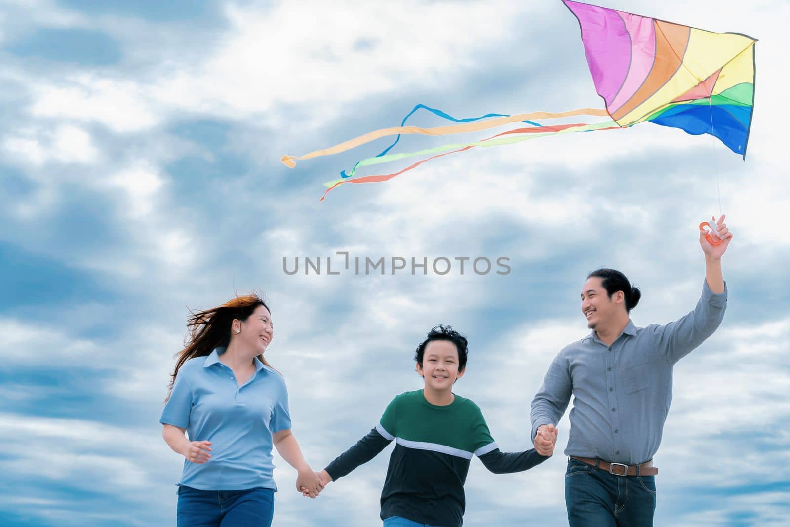 Progressive happy family vacation and carefree day concept. Young parents mother father and son run along and flying kite together road with enjoy natural scenic on scenery and clear sky background.