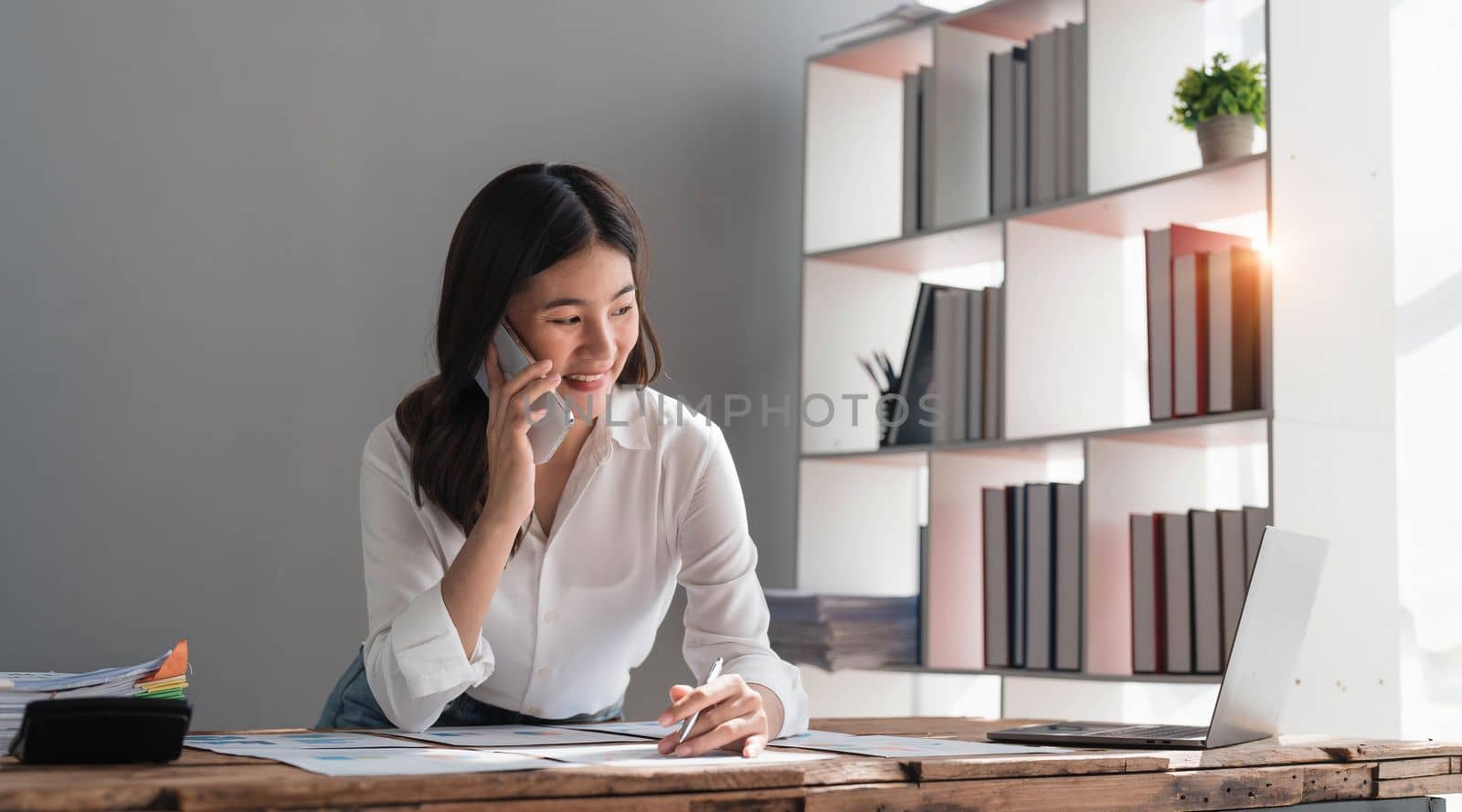 Beautiful Asian businesswoman is using smartphone to chat with customers at work and play various social media applications in a relaxed smile and good mood. by wichayada