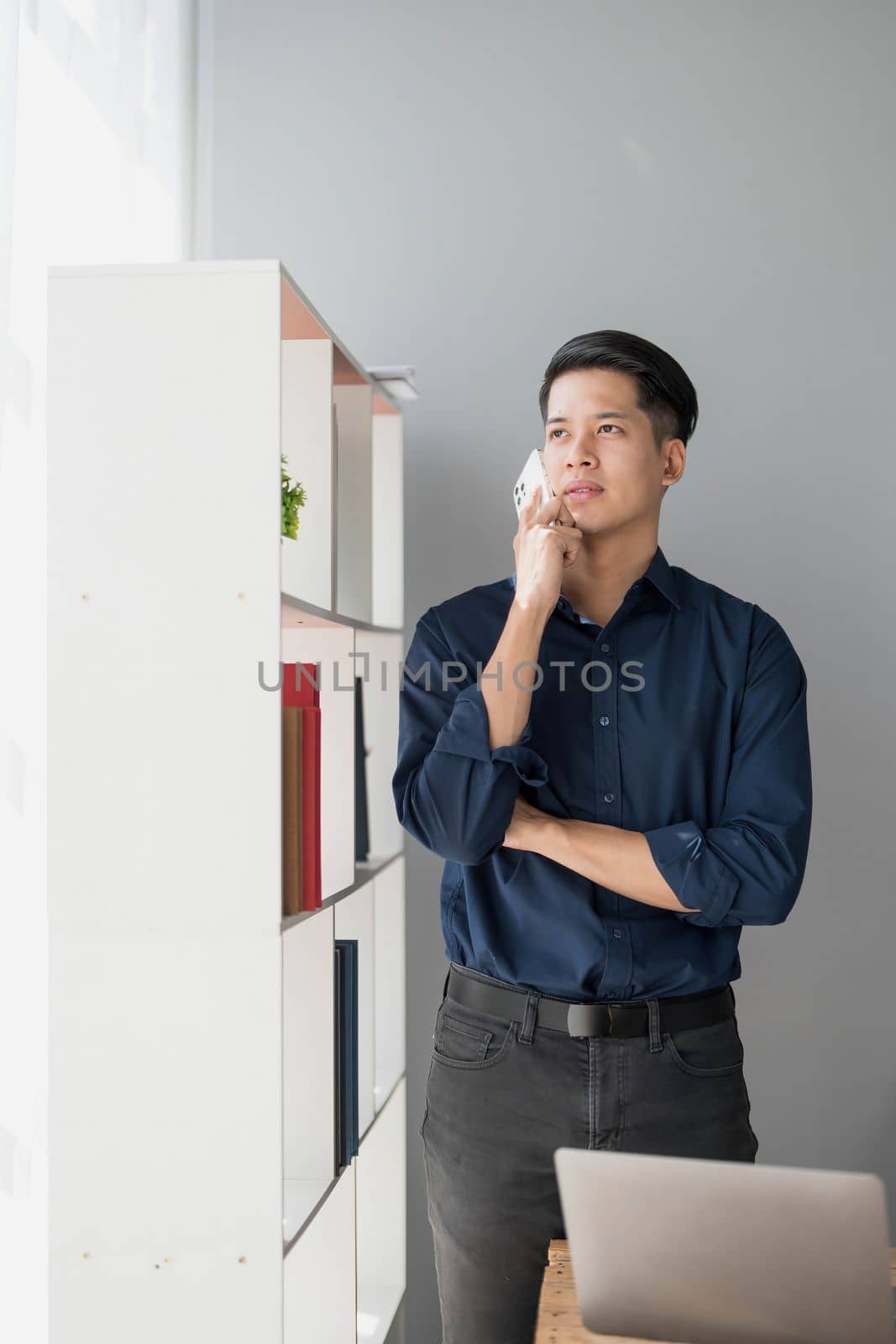 A young Asian businessman stands on the office of a window talking on the phone...